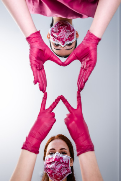 Zwei Frauen mit gemusterten Masken und pinken Handschuhen formen gemeinsam mit ihren Händen ein Herzsymbol in Richtung Kamera.